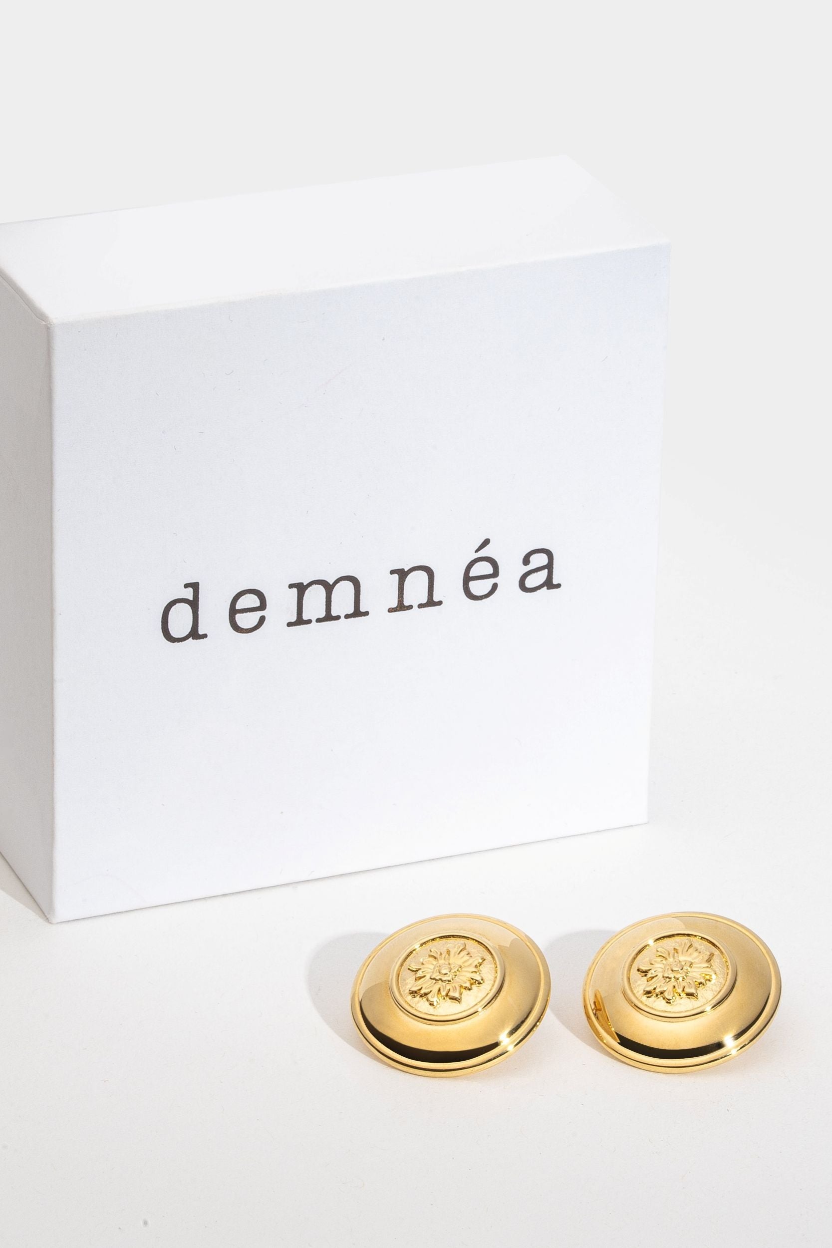 Gold earrings with 'demneéa' branding on a white background