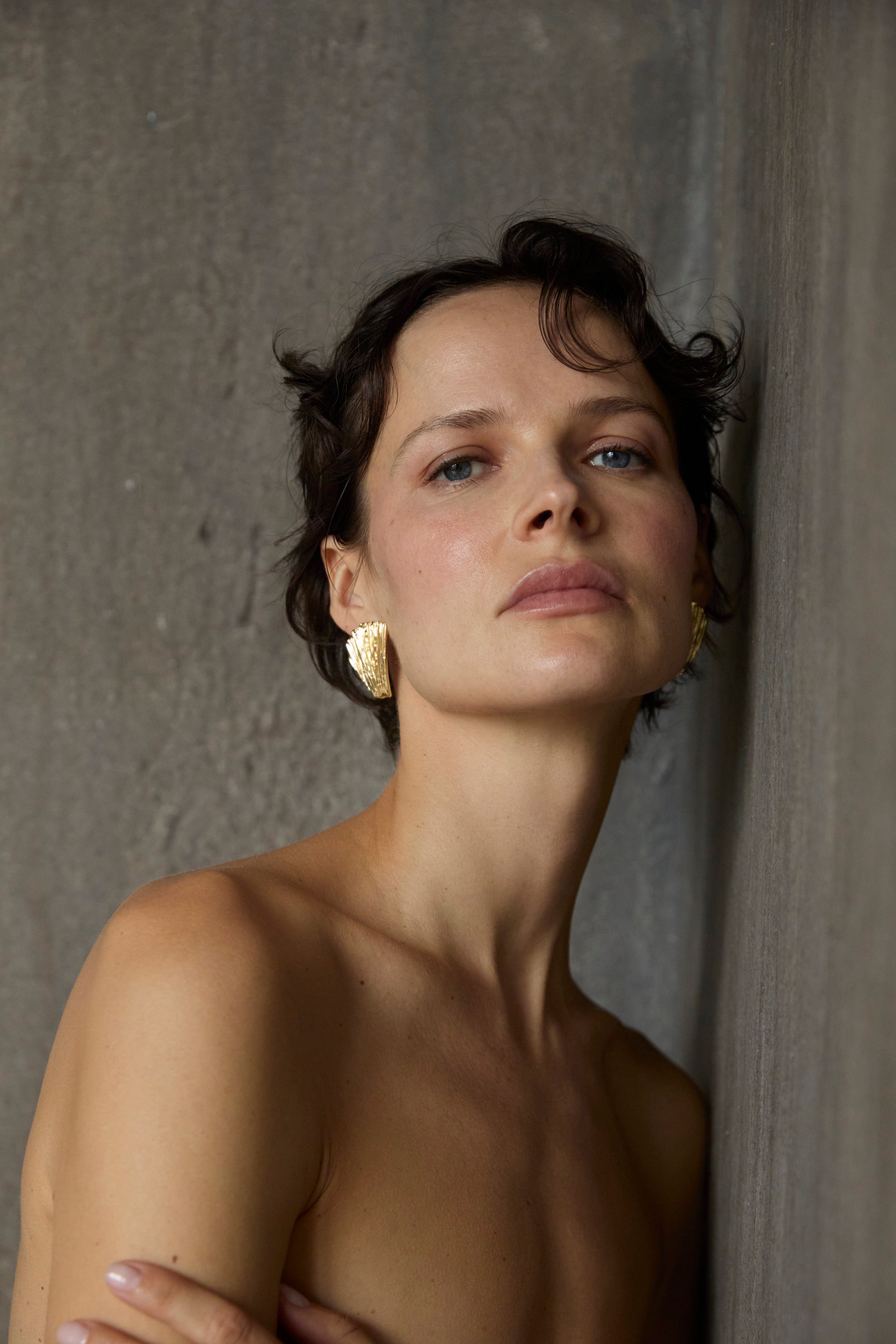 Naked woman leaning against a textured wall with Neréa earrings on